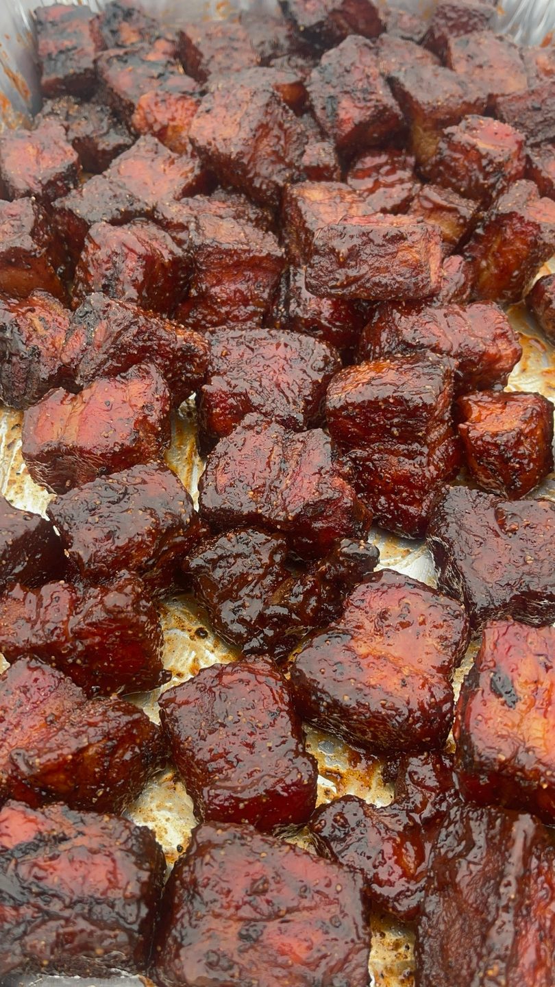 Close-up of numerous glistening, dark brown, square-cut pieces of barbecue meat in a pan.