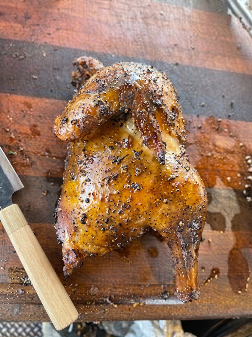 A whole roasted chicken, glistening with sauce and black pepper, rests on a wooden cutting board next to a knife.